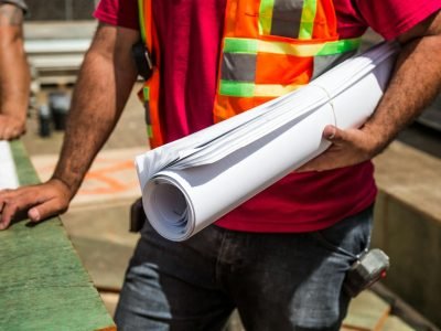 Construction workers in safety vests holding blueprints at a site during the day.
