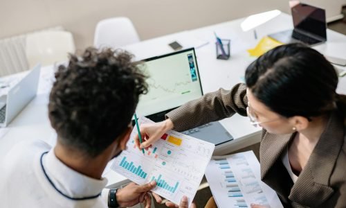 Two colleagues in a meeting room discussing financial charts and graphs on a laptop and paper.