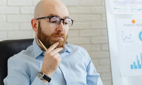 Bearded man in blue shirt contemplating financial data on whiteboard indoors.